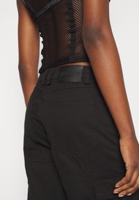 Personne portant un haut en maille noire et un pantalon noir avec une poche arrière et des passants de ceinture, main reposant près de la hanche.