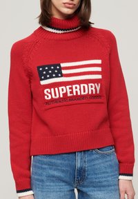 Rode gebreide trui met hoge kraag, voorzien van een witte en blauwe Amerikaanse vlag graphic en "SUPERDRY" tekst. Geribde boorden en zoom.