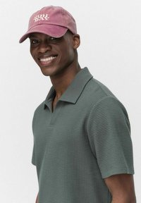 Mörkgrön kortärmad pikétröja med våffeltextur, kombinerad med en rosa keps med vit text "SUN DAY".