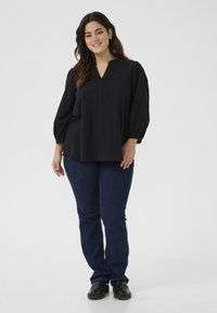 Schwarzes, plissiertes Oberteil mit V-Ausschnitt und Puffärmeln, kombiniert mit dunkelblauen Jeans und schwarzen Schuhen. Glattes Material und lockere Passform.