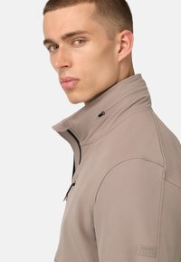 Veste zippée beige clair avec col haut, accents de zip noirs et finition texturée. Présente un logo discret sur la manche.
