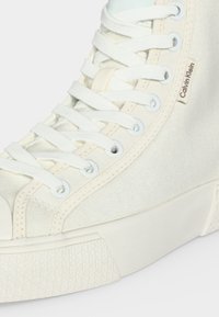 Gros plan d'une basket montante en toile blanche avec des lacets blancs et une étiquette logo Calvin Klein sur le côté près de la cheville.