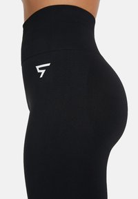 Crne tajice visoki struk od glatkog materijala sa bijelim logotipom na donjoj strani, s konturiranim krojem koji ističe prirodne obline.