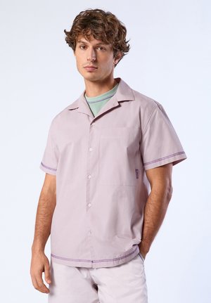 Jeune homme portant une chemise à manches courtes boutonnée mauve clair par-dessus un t-shirt vert à col rond, se tenant devant un fond clair uni.