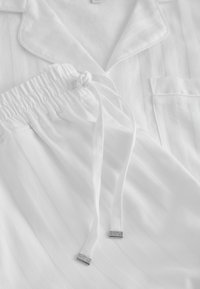 Ensemble de pyjama en coton blanc avec une chemise à col boutonné et un pantalon à taille élastique avec des cordons noués aux embouts métalliques argentés.