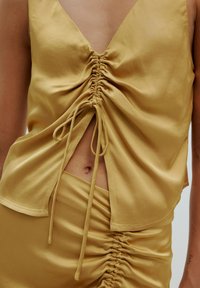 Lichtgele top met diepe V-hals, voorruching en gestrikt detail. Gladde textuur en een vloeiende pasvorm, gecombineerd met een bijpassende geruchte rok.