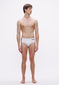 Weiße Badehose mit niedrig geschnittenem Bund, versehen mit einem schwarzen Logo und mehrfarbigen Seitenakzenten. Glatte Textur, minimalistisches Design.