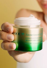 Grön och klar glasburk med Nuxe kräm, med ett vitt lock och präglad text. Krämig konsistens synlig på toppen. Hand som håller burken.