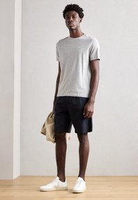 Tricou gri cu mânecă scurtă, pantaloni scurți bleumarin, adidași albi. Pe un fond deschis, ținând o jachetă bej. Design simplu și croială casual.