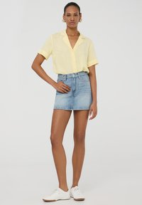 Camicia gialla a maniche corte con bottoni, abbinata a pantaloni corti in denim a vita alta. Modella che indossa sneaker bianche, in piedi su uno sfondo neutro.