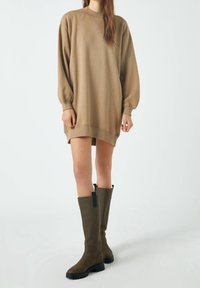 Osoba ubrana w luźną beżową sukienkę swetrową z długim rękawem, w parze z oliwkowymi kozakami do kolan, stojąca na jednolitym jasnym tle.