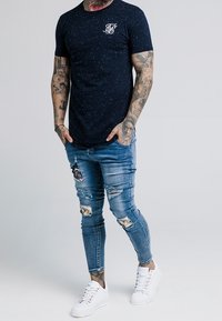 Námořnické tričko s krátkým rukávem a skvrnitým vzorem, bílé logo na hrudi, spojené s prodřenými modrými skinny džíny a bílými teniskami.