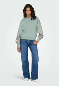 Gaiši zaļš sporta krekls ar ziedu raksta piedurknēm, apvienots ar plašām, zilu džinsu biksēm un baltām kurpēm. Mīksta auduma un brīva piegriezuma.