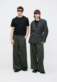 Μαύρο μπλουζάκι, χακί παντελόνι φαρδύ σε ανδρικό μοντέλο· γκρι διπλό σακάκι με ζώνη σε γυναικείο μοντέλο. Και οι δύο φορούν μαύρα παπούτσια.