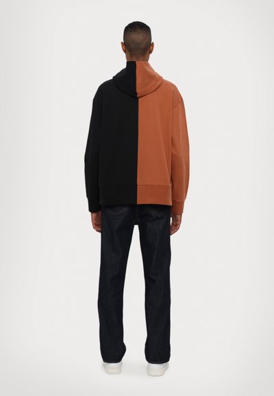 Farblich abgesetzter Hoodie mit schwarzen und braunen Abschnitten, einem geräumigen Kapuzenbereich und gerippten Bündchen, kombiniert mit dunklen Jeans und weißen Sneakers.