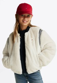 Biała kurtka bomberka z sztucznego futra z czarnymi akcentami żebrowanymi, z zamkiem z przodu i granatowym swetrem pod spodem. Czerwona czapka baseballowa dopełnia wygląd.