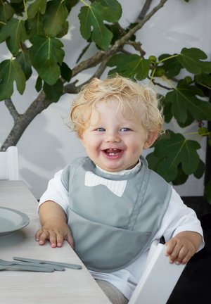 Bavaglino in silicone grigio con un design a farfallino bianco, bambino con capelli biondi ricci, seduto a un tavolo con posate e piatti grigi. Foliage verde sullo sfondo.