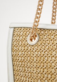 Torebka z plecionej słomy z teksturowanym beżowym wykończeniem, białymi detalami i złotymi łańcuszkami jako uchwytami, z okrągłym otworem w detalach.
