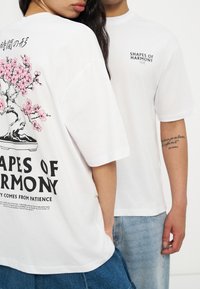 Δύο άτομα φορούν λευκές μπλούζες με το κείμενο "Shapes of Harmony"· το ένα δείχνει ένα ροζ γραφικό κερασιάς στην πλάτη.