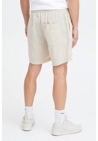 Pantaloni scurți bej, cu textură, având o talie elastică, un buzunar în spate și o croială lejeră, asortați cu adidași și șosete albe.