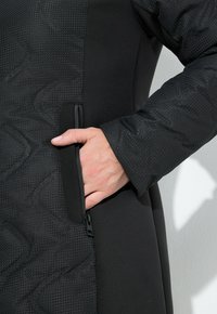 En hand som för in i en blixtlåsficka på ärmen av en svart, texturerad och slät tygjacka mot en enfärgad bakgrund.