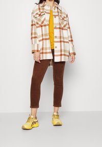 Rutigt jacka i vitt, beige och orange, tillsammans med en gul topp och bruna manchesterbyxor. Gula sneakers fulländar outfiten.