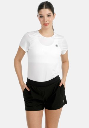 Frau mit dunklem Haar im Pferdeschwanz, die ein weißes kurzärmeliges Shirt und schwarze Shorts trägt und mit den Händen in den Taschen vor weißem Hintergrund steht.