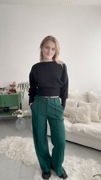 Νεαρή γυναίκα που στέκεται σε εσωτερικό χώρο, φορώντας μαύρη μπλούζα με μακριά μανίκια και πράσινο παντελόνι με φαρδιά μπατζάκια, με τα χέρια στις τσέπες, χαμογελώντας απαλά.