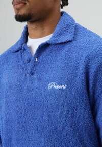 Blauw polohemd met een zachte, pluizige textuur, inclusief een kraag, een twee-knopenbies en een geborduurd "Present"-logo op de borst. Zacht materiaal, casual ontwerp.