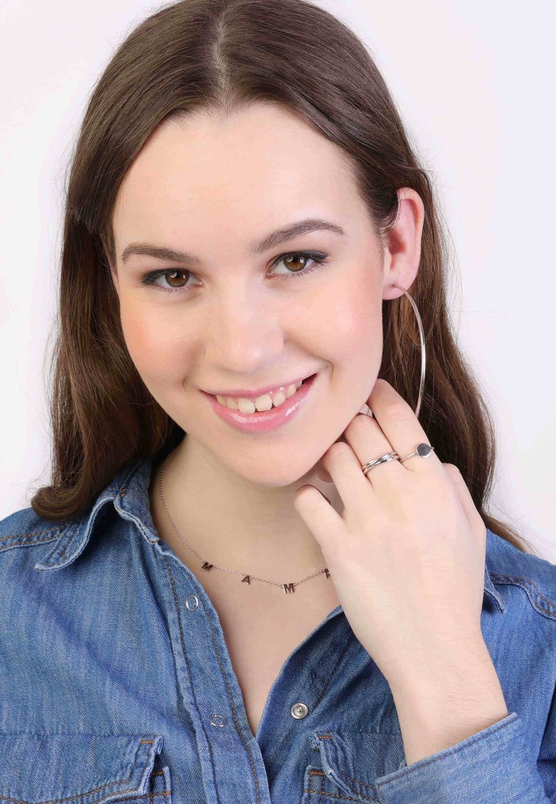 Zilveren hoopsieraden, een delicate ketting met letteraccenten die "MAMA" spellen, en eenvoudige gestapelde ringen tegen een achtergrond van een denim overhemd.