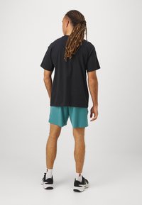 Camiseta negra de manga corta combinada con shorts en color teal. El modelo lleva zapatillas negras y blancas con acentos de malla visibles.