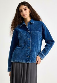 Tmavě modrá džínová bunda s vybledlou texturou, vybavená dvěma předními kapsami, zapínáním na knoflíky a kontrastním prošitím podél okrajů.