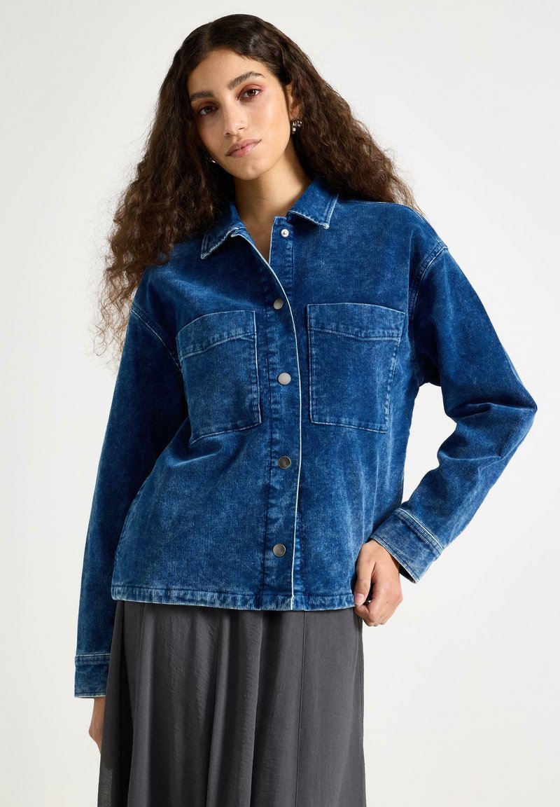 Tmavě modrá džínová bunda s vybledlou texturou, vybavená dvěma předními kapsami, zapínáním na knoflíky a kontrastním prošitím podél okrajů.