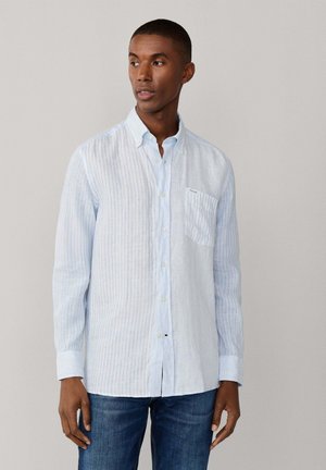 Hombre con camisa de botones de rayas verticales blancas y azul claro de manga larga y vaqueros azules, mirando hacia su izquierda.