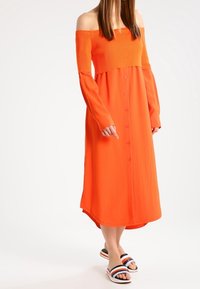 Femme portant une robe midi orange longue à manches longues et épaules dénudées, avec des boutons à l'avant et des sandales à enfiler ouvertes à rayures.