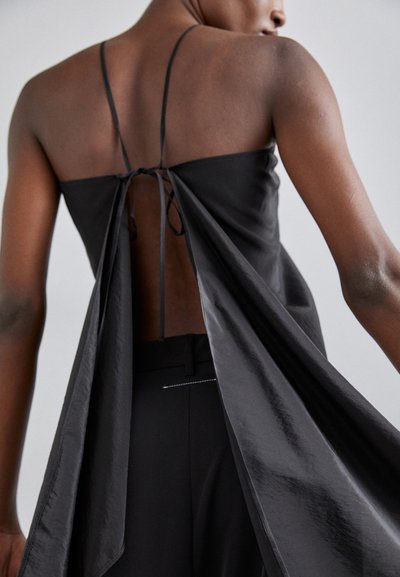 Black sleeveless top with a tied back, featuring a flowing draped design. Smooth texture and minimalist style, paired with black pants.