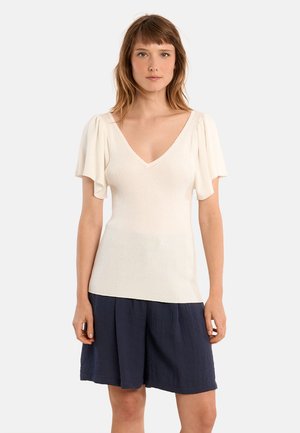 Mujer con una blusa de canalé blanca de cuello en V y mangas con volantes, y pantalones cortos plisados de color azul marino oscuro, de pie frente a un fondo liso.