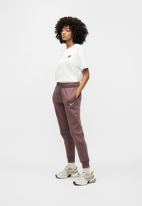 Λευκό κοντομάνικο μπλουζάκι με μαύρο λογότυπο Nike, συνδυασμένο με μωβ φόρμες με ελαστική μέση και πλαϊνές τσέπες. Μπεζ αθλητικά παπούτσια.