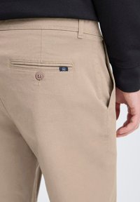 Pantaloni in cotone beige con una tasca posteriore dotata di chiusura a bottone e un piccolo etichetta blu. La texture è liscia, con cuciture pulite.