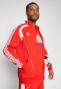 Rød zip-up jakke med hvite striper og logo, båret over en matchende skjorte. Modellen holder en svart og hvit fotball.