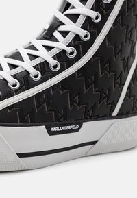 Černá teniska s texturovaným svrškem s opakujícím se vzorem loga, bílými tkaničkami a kontrastní gumovou podrážkou s výrazem značky.