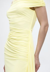 Γυναίκα που φοράει ένα εφαρμοστό ανοιχτό κίτρινο φόρεμα με ρελιασμένες λεπτομέρειες στην πλευρά και κοντά μαζεμένα μανίκια.