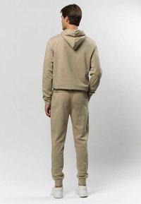 Beige kapucnis pulóver és azonos színű jogging nadrág, amelyek puha anyagból készültek. Húzószerkezetes kapucni és bordázott mandzsetták jellemzik, amelyeket fehér sportcipőkkel viselnek.