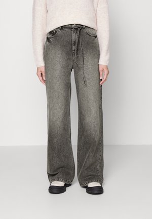 Persoon draagt wijd uitlopende, vervaagde grijze spijkerbroek met een trekkoord in de taille, lichtroze trui en zwart-witte schoenen, staand op een grijze vloer.