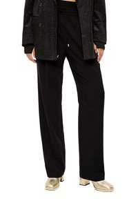 Zwarte wijde broek met een elastische tailleband en zijzakken, gecombineerd met een gestructureerde zwarte jas en gouden hakken.