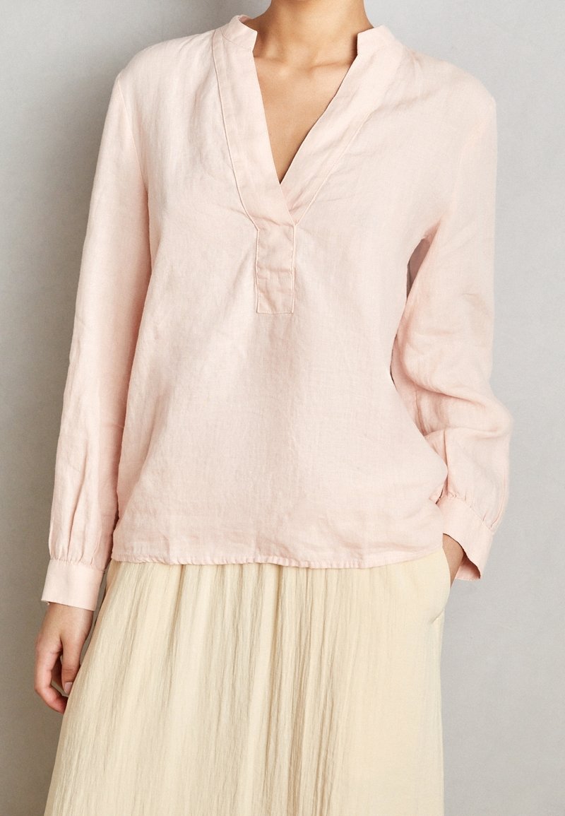 Lichtroze linnen blouse met een v-hals, lange mouwen en subtiele plooidetails bij de halslijn. Zachte textuur, relaxte pasvorm.