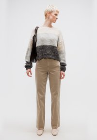 Άτομο με κοντά ξανθά μαλλιά που φοράει ριγέ πλεκτή μπλούζα, μπεζ παντελόνι, κρεμ παπούτσια και κρατάει μια σκουρόχρωμη τσάντα ώμου, στέκεται μπροστά σε λευκό φόντο.