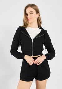 Čierna skrátená mikina s dlhým rukávom a zipsom, s prednou kapsou, skombinovaná s čiernymi šortkami s elastickým pásom. Bavlnený materiál s hladkou štruktúrou.