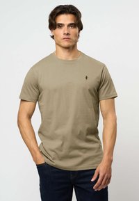 Oliivinvihreä puuvillainen t-paita, jossa on pyöreä kaula-aukko ja lyhyet hihat, ja joka sisältää pienen, tumman logon vasemmassa rinnassa. Yhdistettynä tummiin farkkuihin.