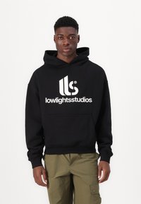 Felpa nera con un grande logo bianco "lowlightsstudios" sul davanti, dotata di tasca frontale e cappuccio con cordino. Indossata con pantaloni cargo verde oliva.